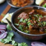 A bowl of spicy Chicken Kalia curry garnished with fresh cilantro, surrounded by sliced red onions, cilantro leaves, and other condiments on a wooden board.