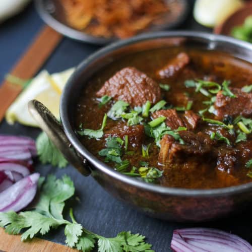 A bowl of spicy Chicken Kalia curry garnished with fresh cilantro, surrounded by sliced red onions, cilantro leaves, and other condiments on a wooden board.