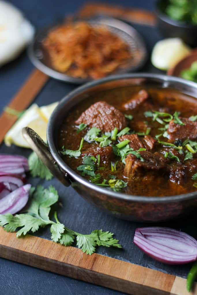 A bowl of spicy Chicken Kalia curry garnished with fresh cilantro, surrounded by sliced red onions, cilantro leaves, and other condiments on a wooden board.