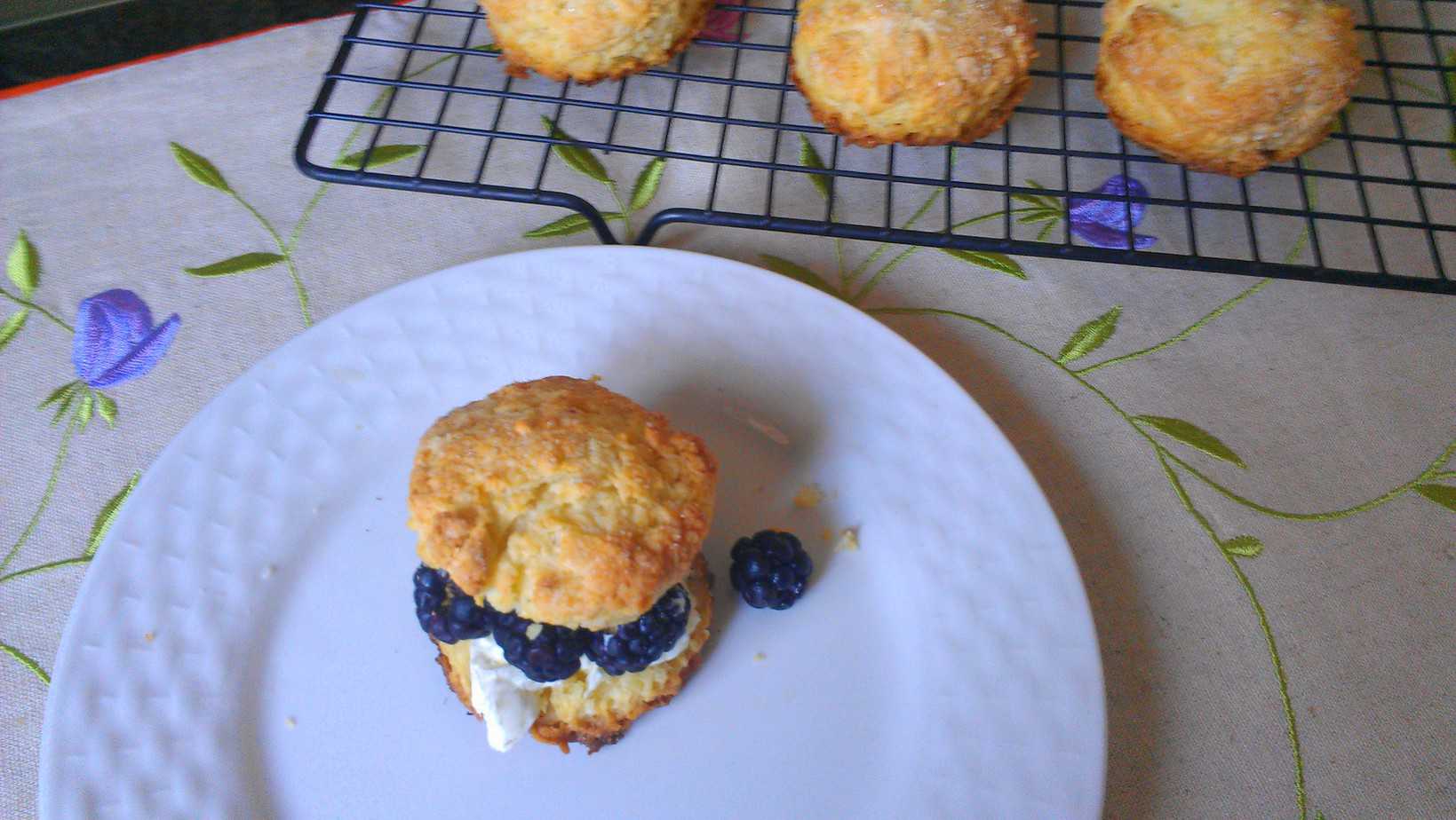 A scone sandwich with fresh blackberries and cream on a white plate, with more scones cooling on a wire rack in the background, atop a tablecloth with floral embroidery.