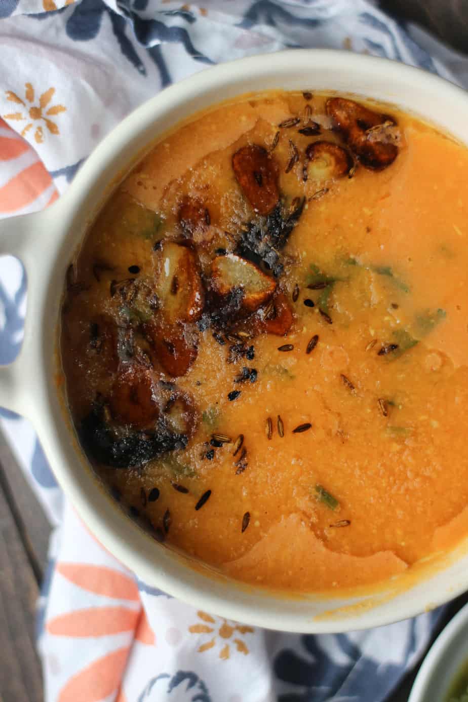 A close-up of a bowl of creamy Masoor ki Daal (orange lentil soup) garnished with browned garlic slices, cumin seeds, and fresh herbs, set on a patterned cloth.
