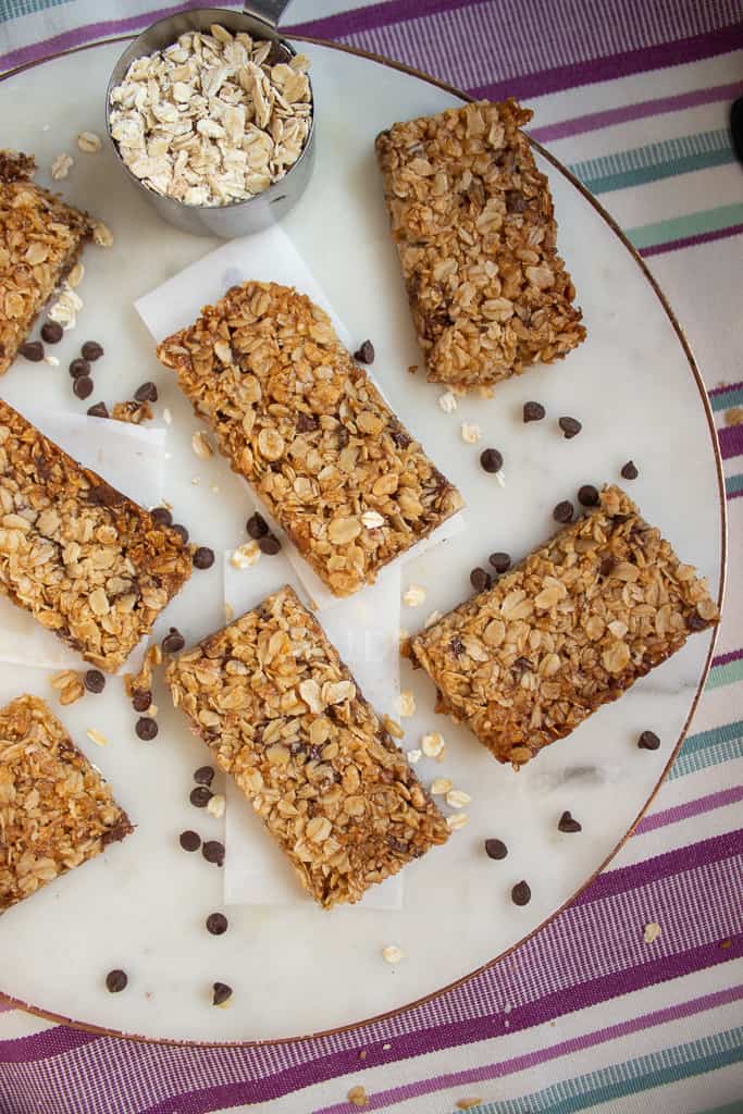 Crunchy Nut Free Granola Bars with oats and chocolate chips are arranged on a white plate. A metal measuring cup filled with oats and scattered chocolate chips add to the scene. The plate sits atop a striped, colorful cloth.