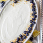 An oval white dish filled with easy lemon mousse, topped with a border of fresh blueberries and sprinkled graham cracker crumbs. Lemon wedges and extra blueberries are scattered on a silver tray underneath.