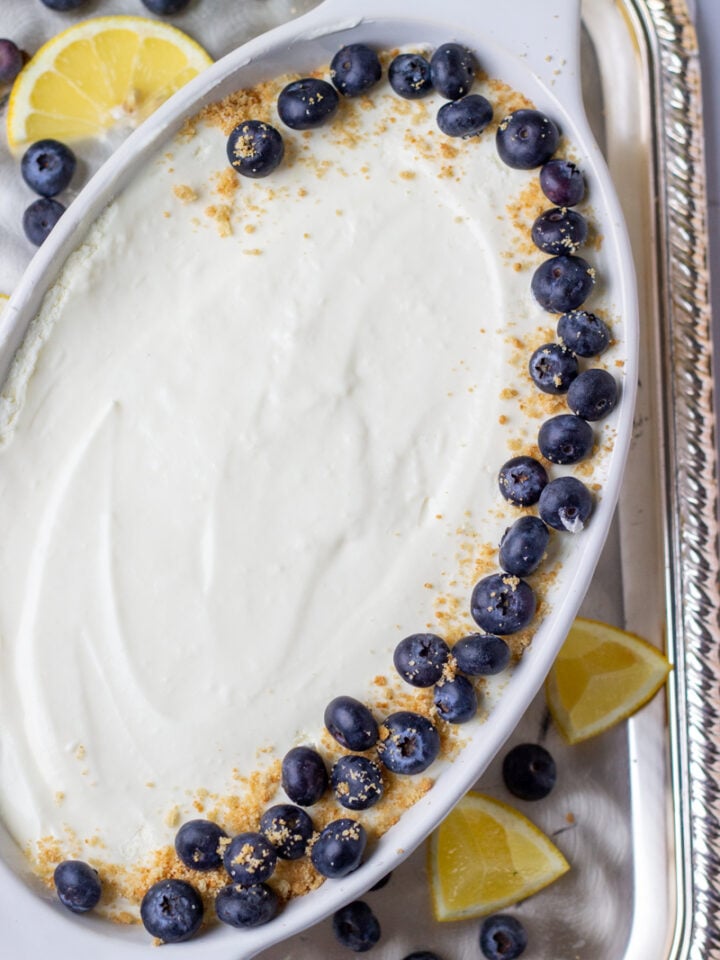 An oval white dish filled with easy lemon mousse, topped with a border of fresh blueberries and sprinkled graham cracker crumbs. Lemon wedges and extra blueberries are scattered on a silver tray underneath.