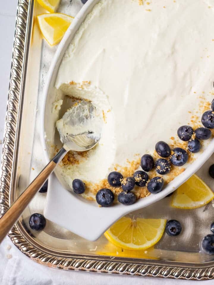 An oval white dish filled with easy lemon mousse, topped with a border of fresh blueberries and sprinkled graham cracker crumbs. Lemon wedges and extra blueberries are scattered on a silver tray underneath.