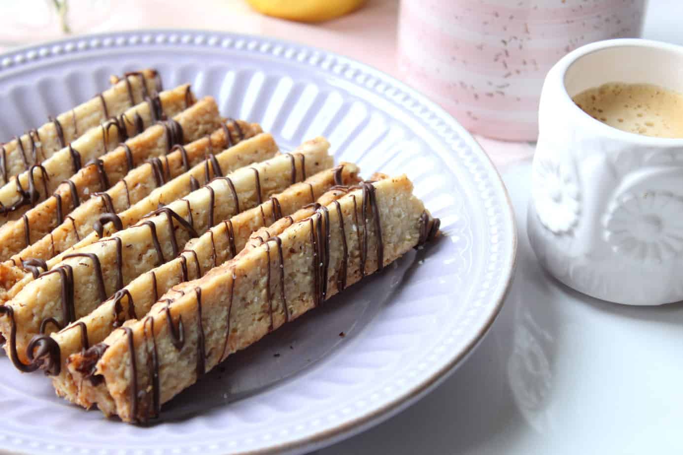 A lavender plate holds several almond cookies drizzled with chocolate, arranged in neat rows beside some Crunchy Hazelnut Sticks. Next to the plate is a white cup filled with espresso.