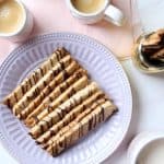 A plate of crunchy hazelnut sticks drizzled with chocolate sits on a lavender plate, surrounded by cups of coffee, a jar with more biscotti, and a pink napkin on a white table.