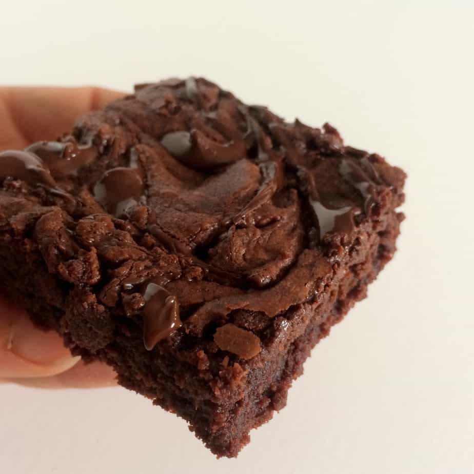 A hand holding a thick, fudgy Ferrero Rocher Brownie with visible chocolate chips on top, against a plain white background.