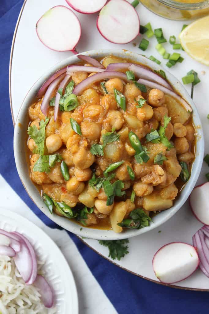 A bowl of aloo chana curry garnished with chopped onions, green chilies, and cilantro, surrounded by sliced radishes, green onions, lemon wedge, and a plate of rice on the side.
