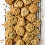 A cooling rack filled with a pile of chocolate chip cookies and Butterscotch Chip Cookies rests on a white surface, with a few scattered chips all around.