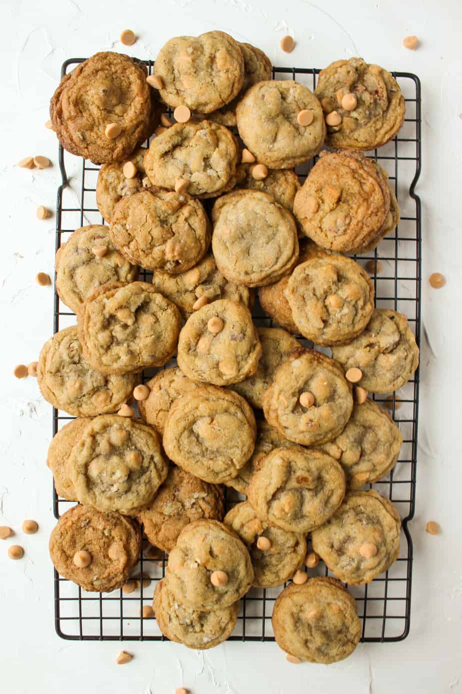 A cooling rack filled with a pile of chocolate chip cookies and Butterscotch Chip Cookies rests on a white surface, with a few scattered chips all around.