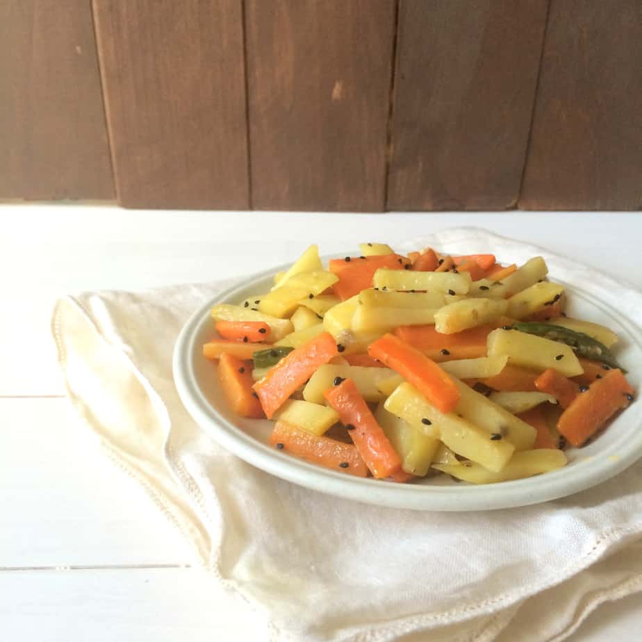 A white plate filled with sautéed carrot and potato sticks, known as Aaloo Gajar, garnished with black sesame seeds, placed on a folded white cloth atop a white wooden surface with a wooden wall in the background.