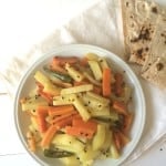 A white plate with stir-fried Aaloo Gajar—spicy carrots and potatoes—garnished with black sesame seeds, sits next to folded pieces of flatbread on a white napkin atop a light-colored table.