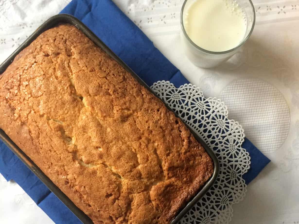 A golden-brown cream cheese pound cake in a pan rests on a blue cloth, next to a glass of milk and a decorative white doily on a white tablecloth.