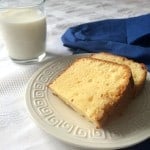 Two slices of cream cheese pound cake on a white plate with a geometric pattern, beside a glass of milk and a blue napkin atop a white tablecloth.