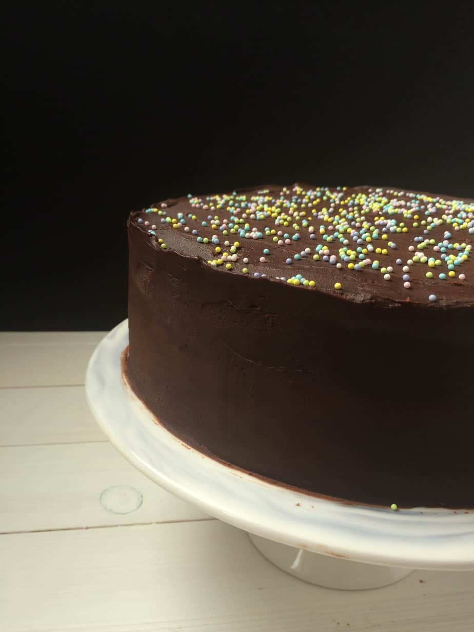 A One Bowl Chocoholics Chocolate Cake, frosted with rich chocolate and topped with colorful round sprinkles, sits on a white cake stand against a black background and light wooden surface.