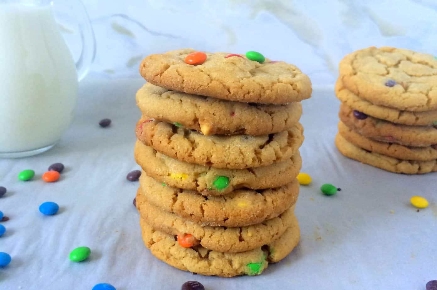 A stack of colorful sweet and salty peanut butter cookies is arranged on a white surface, with scattered candies and a glass pitcher of milk in the background. Another stack of cookies is visible to the right.