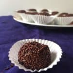 A close-up of a brigadeiro chocolate truffle covered in chocolate sprinkles, placed in a white paper cup on a dark blue cloth, with more brigadeiros in white cups on a plate in the background.