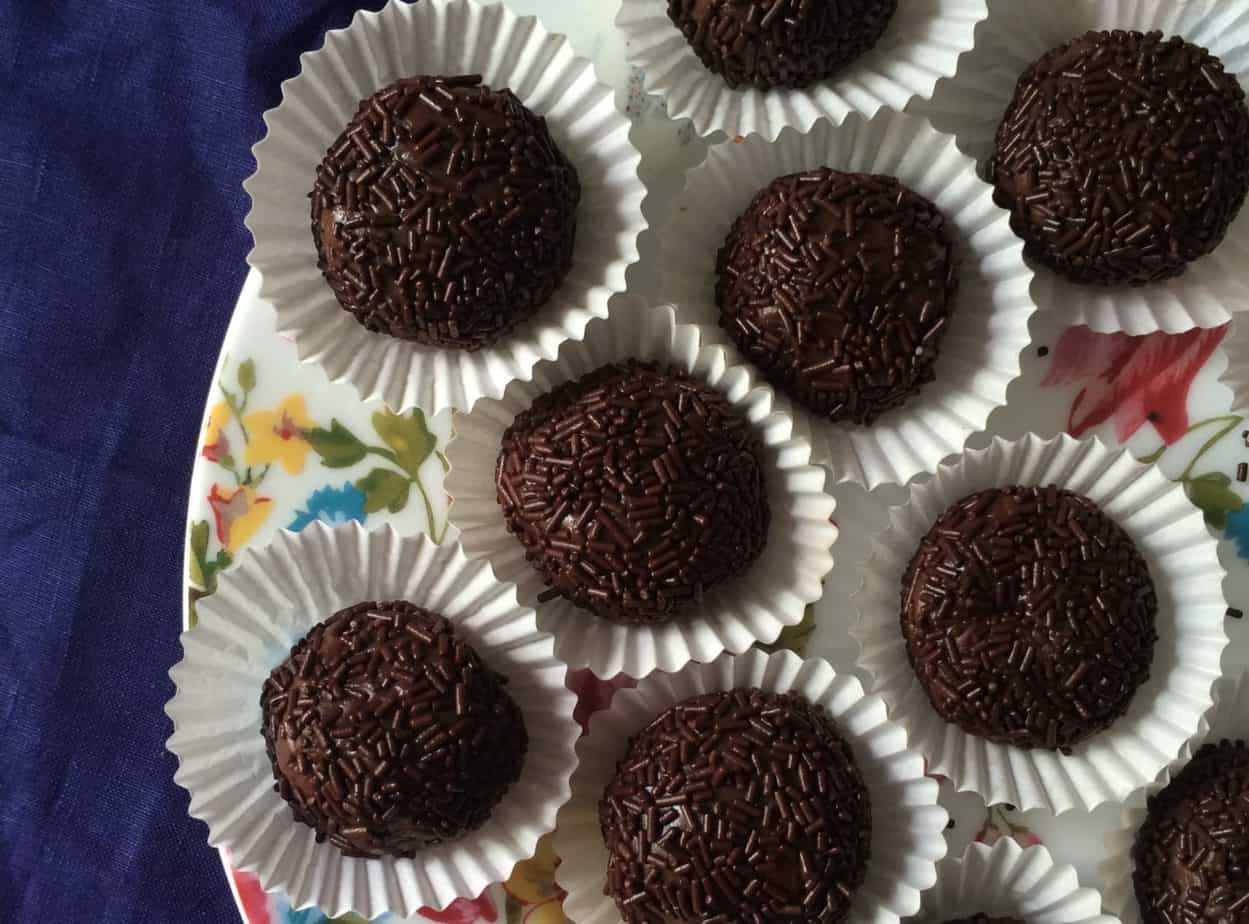 Brigadeiros coated with chocolate sprinkles are arranged in white paper cups on a floral-patterned plate, with a dark blue cloth partially visible beside the plate.