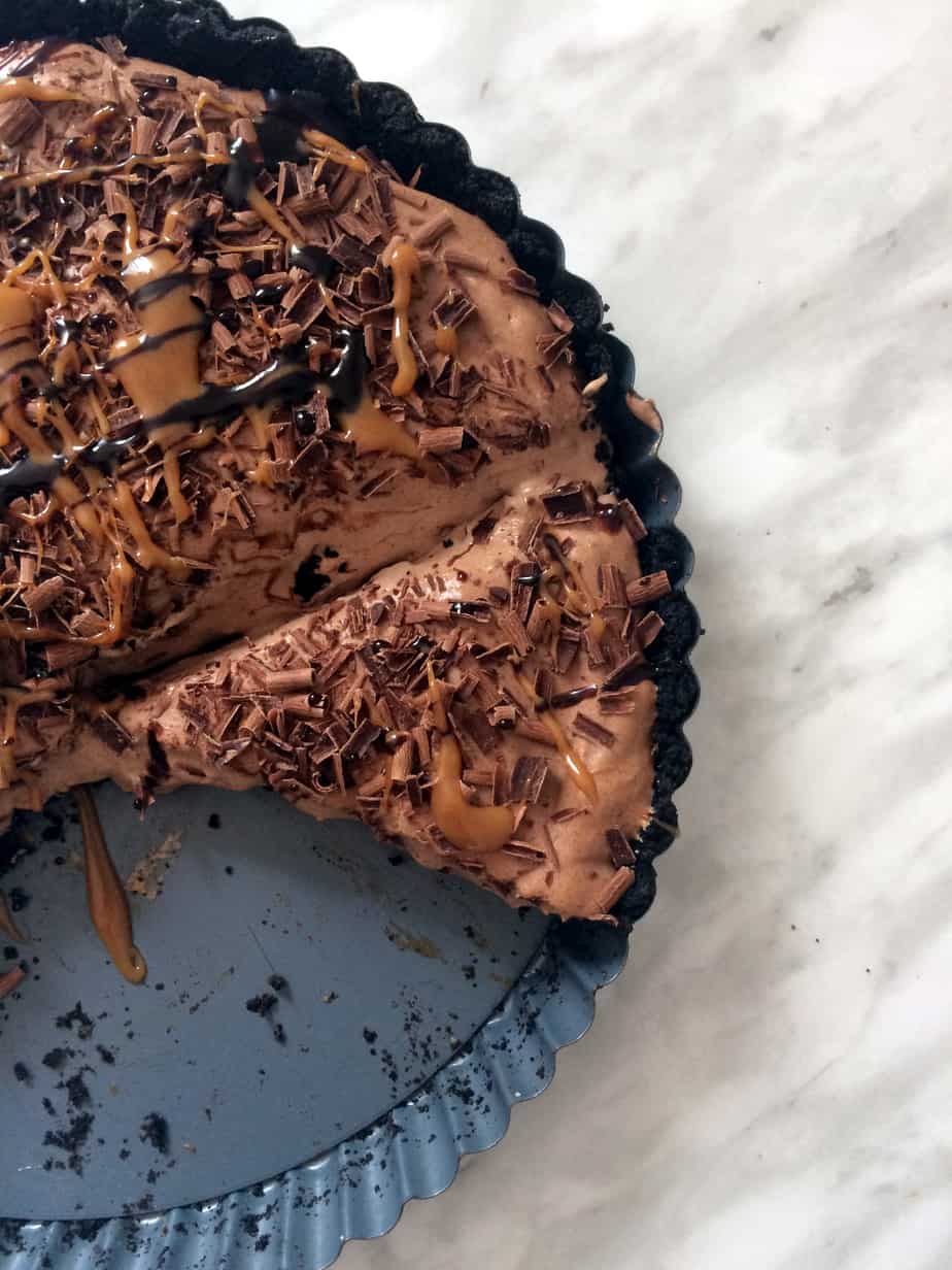 A chocolate mousse tart with a slice missing, topped with chocolate shavings, chocolate drizzle, and caramel sauce, sits in a dark pan on a marble surface.