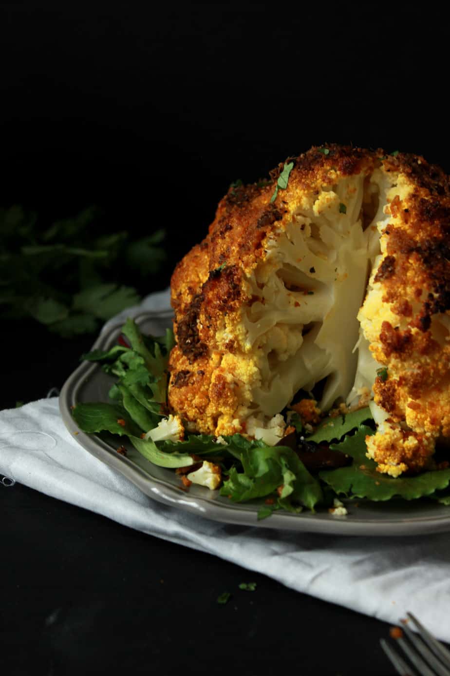 A Spicy Whole Roasted Cauliflower with a golden crust sits on a bed of leafy greens, partially cut open to reveal the tender inside, against a dark background.