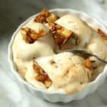 A small white bowl filled with creamy ice cream sits on a marble surface, topped with caramel crunch clusters and a spoon resting in the bowl.