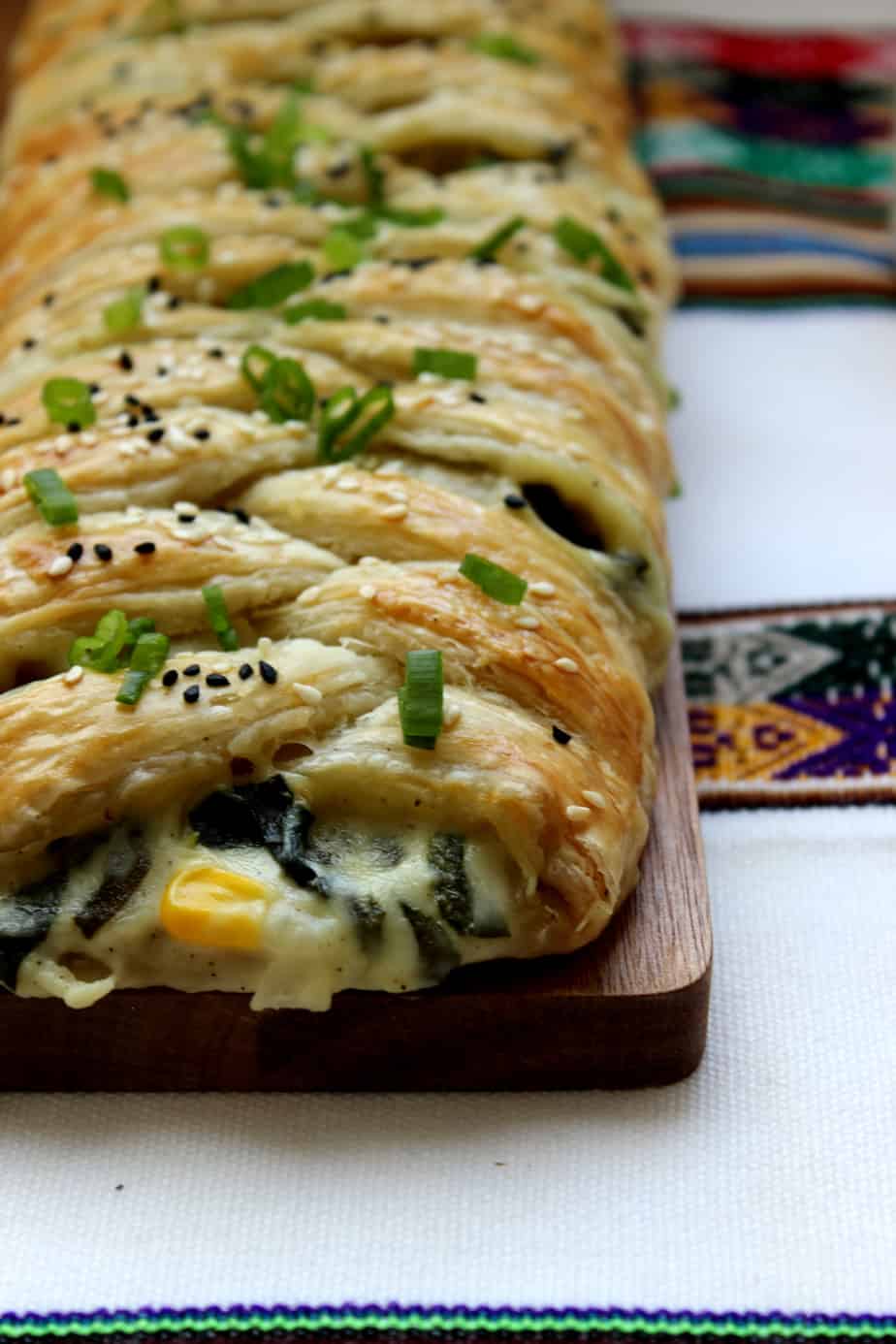 A close-up of a golden-brown puff pastry braid filled with cheese and spinach, topped with chopped green onions and black sesame seeds, resting on a wooden board with colorful woven fabric in the background.