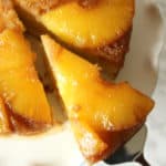A close-up of a Pineapple Upside Down Cake on a white plate. One slice is being lifted out with a cake server, revealing caramelized pineapple rings topping the golden Pineapple Upside Down Cake.