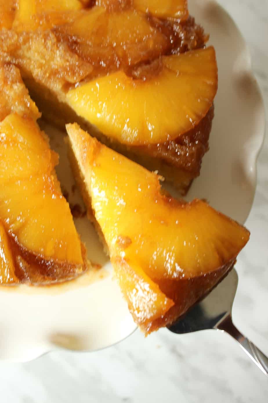 A close-up of a Pineapple Upside Down Cake on a white plate. One slice is being lifted out with a cake server, revealing caramelized pineapple rings topping the golden Pineapple Upside Down Cake.