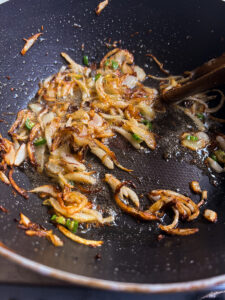 Sliced onions and green chilies being sautéed and caramelized in oil in a black nonstick pan, stirred with a wooden spatula for a flavorful Bhindi Masala Recipe.