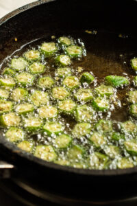 Sliced okra pieces frying in hot oil in a black skillet, with bubbles forming around the okra as it cooks-a delicious start to any Bhindi Masala Recipe.