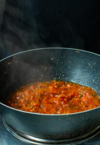A black pan on a stove filled with chunky red tomato sauce simmering, steam rising against a dark background-perfect for starting your Bhindi Masala Recipe.