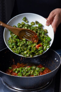 A person pours chopped okra from a white plate into a black pan filled with tomato sauce, using a wooden slotted spoon, preparing the base for a flavorful Bhindi Masala Recipe.