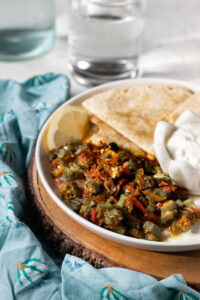 A plate featuring Bhindi Masala Recipe with cooked okra and tomatoes, two pieces of flatbread, a lemon wedge, and a small bowl of yogurt sits on a wooden board, accented by a blue patterned cloth and a glass of water in the background.