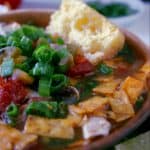 A bowl of Instant Pot Tortilla Soup topped with chopped green onions, diced tomatoes, and crispy tortilla strips, served with a piece of cornbread on the side. A small plate of extra green onions is in the background.