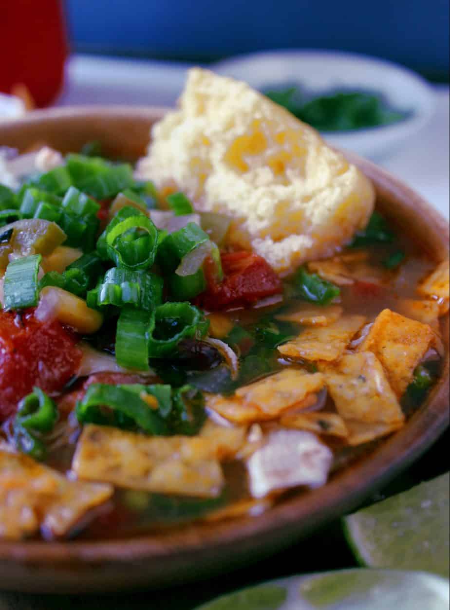 A bowl of Instant Pot Tortilla Soup topped with chopped green onions, diced tomatoes, and crispy tortilla strips, served with a piece of cornbread on the side. A small plate of extra green onions is in the background.