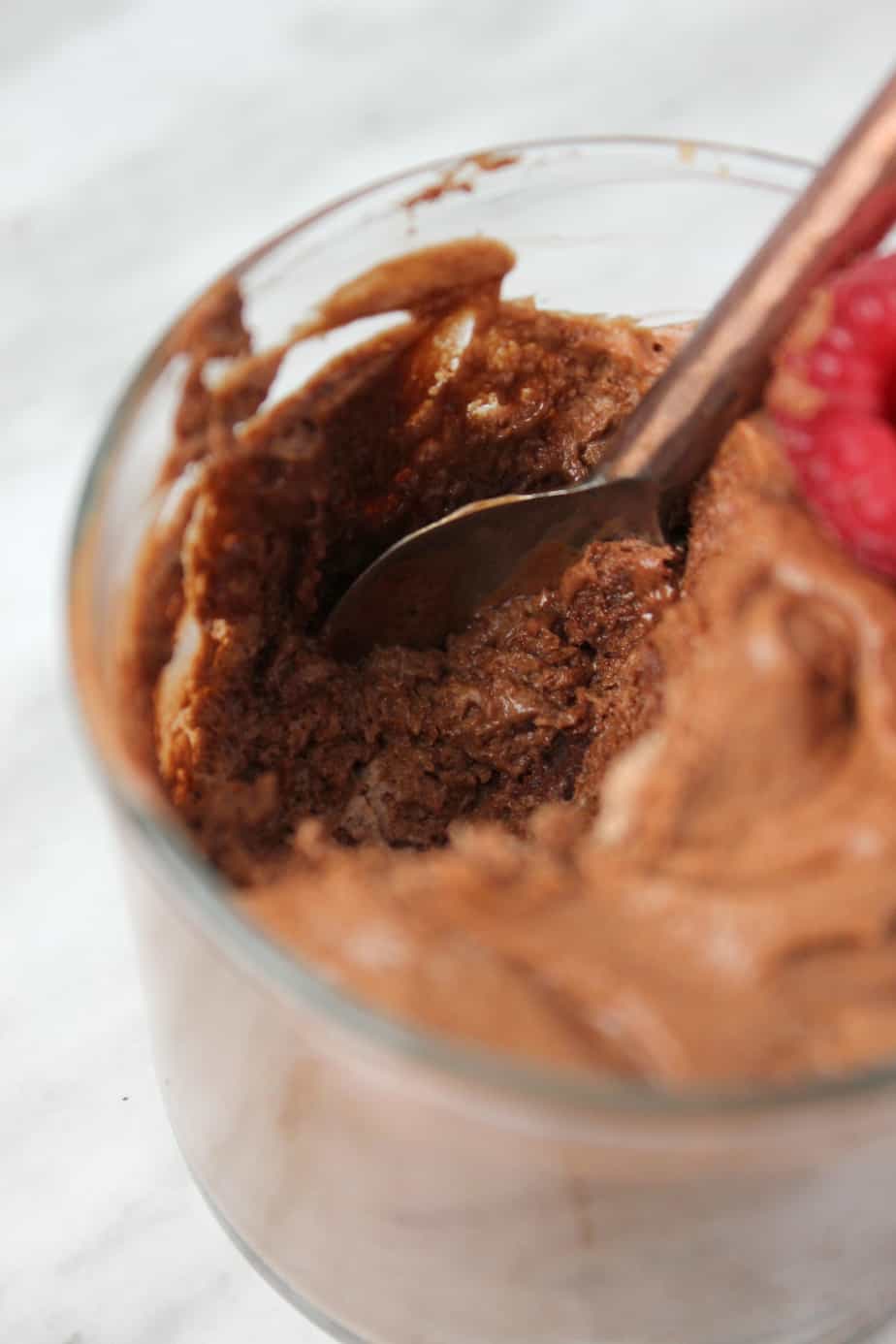 Close-up of a glass filled with creamy chocolate mousse, with a spoon scooping some out. A raspberry sits on top as garnish, highlighting the rich texture of this classic Creamy Chocolate Mousse dessert.