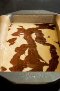 A metal baking pan lined with parchment paper holds unbaked Marble Cake with Oil batter, showcasing beautiful chocolate and vanilla swirls on the surface.