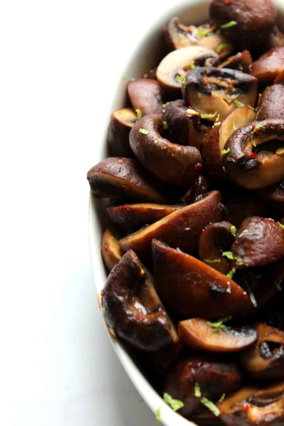 A close-up of a white dish filled with delicious roasted mushrooms, garnished with small bits of green herbs, on a white background.