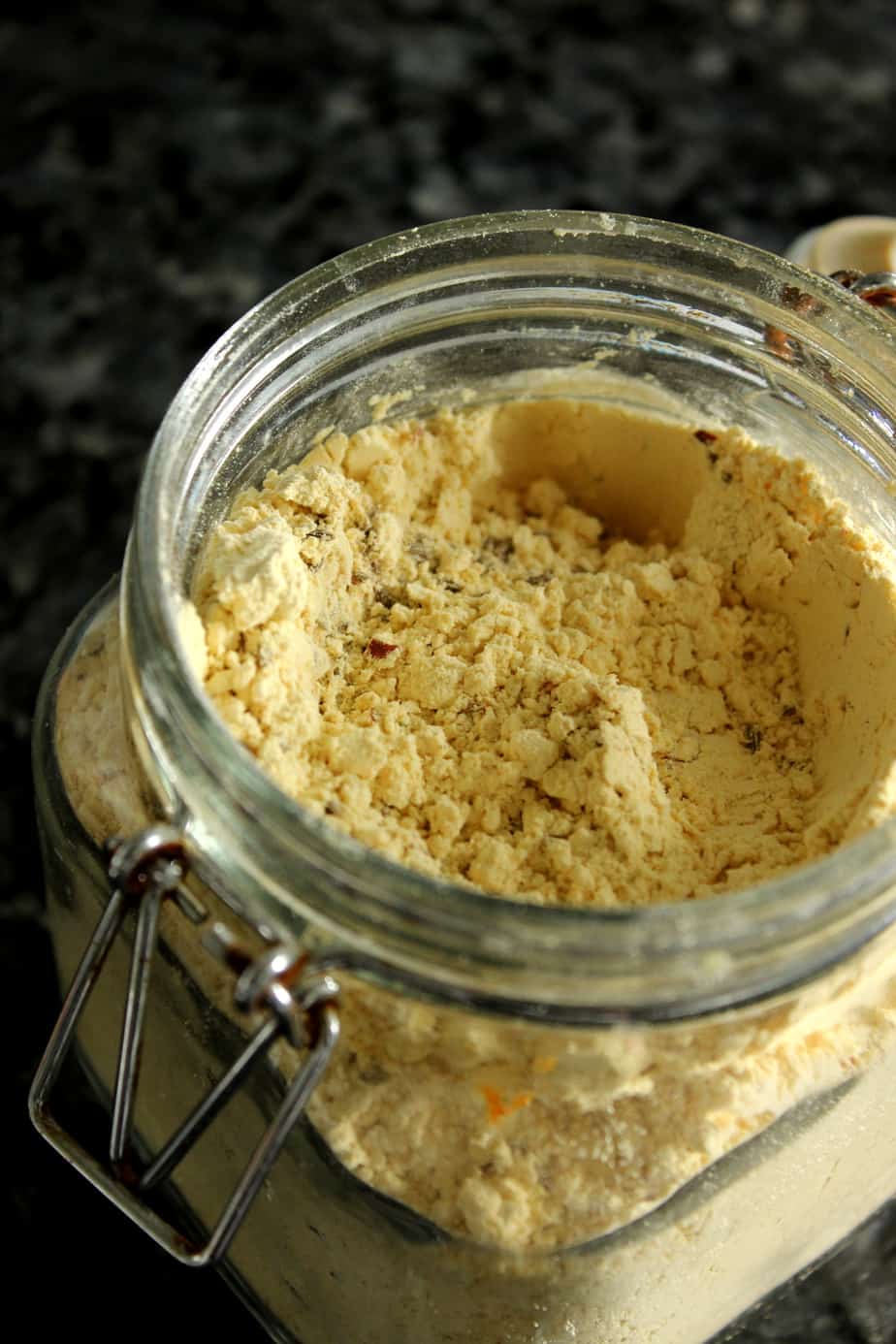 A close-up of an open glass jar filled with a crumbly, light yellow powder-possibly a homemade pakora mix, perfect for anyone learning How to Make Pakoras-sitting on a dark countertop.