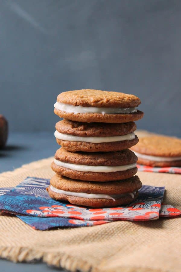 A stack of five sandwich cookies with cream filling sits on colorful napkins atop a burlap cloth, evoking the cozy appeal of Chewy Ginger Cookies, with a blurred cookie and neutral background behind.