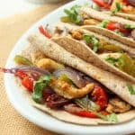 Three tortillas filled with baked chicken fajitas—seasoned chicken strips, red and green bell peppers, and onions—topped with chopped cilantro, served on a white plate. A bowl of salsa and green garnish are blurred in the background.