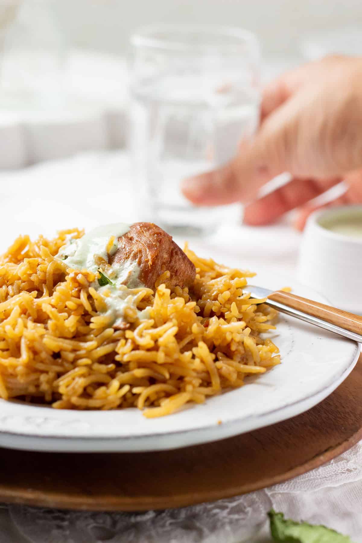 A plate of pakistani chicken pulao drizzled with raita and a hand in the background reaching for a glass.