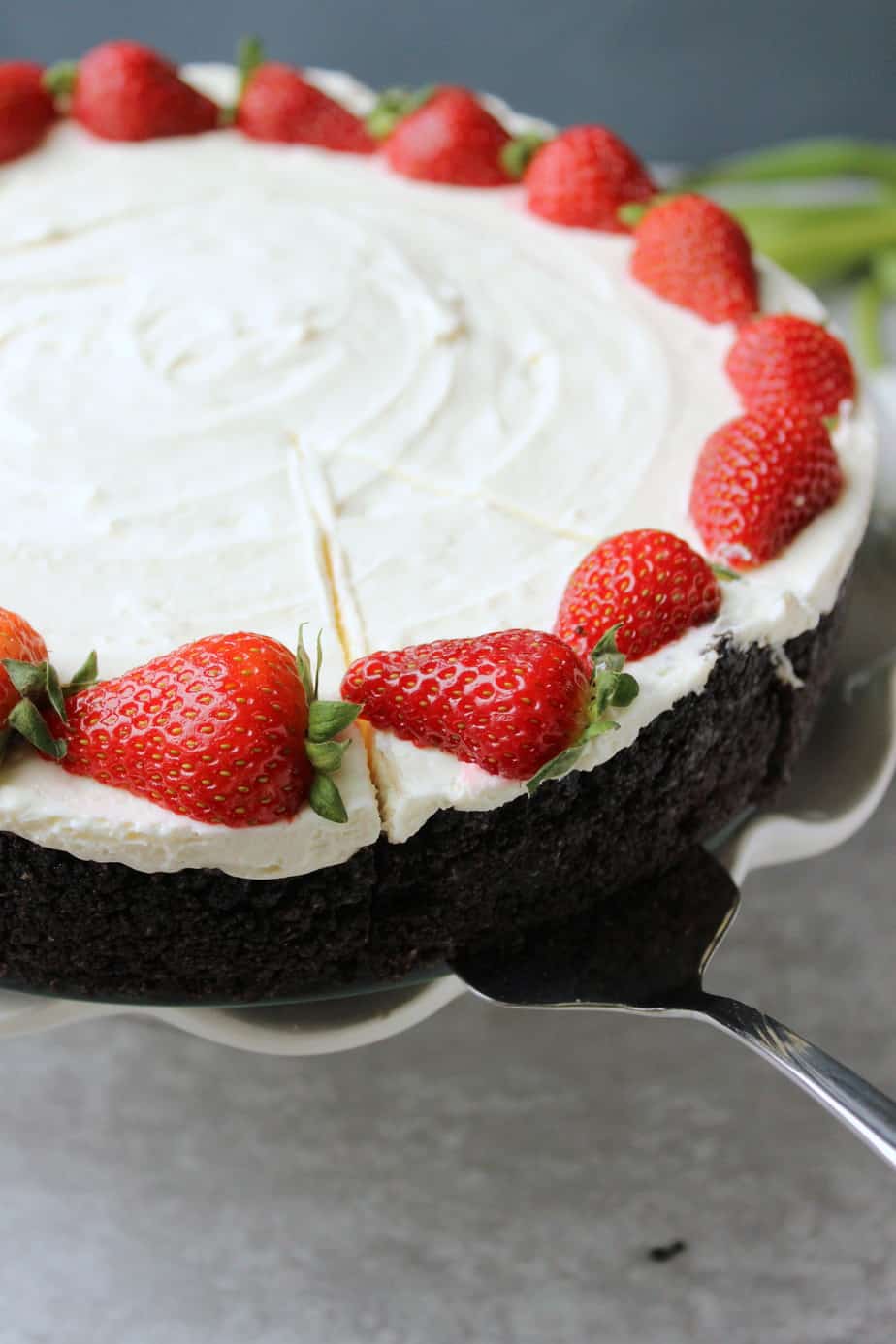 A chocolate cake with white frosting, topped with whole strawberries along the edge, sits on a cake stand. Perfect for celebrations, it offers a tempting look reminiscent of the Best No Bake Oreo Cheesecake. A slice is being lifted with a metal cake server.