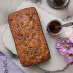 A loaf of banana bread with walnuts rests on a marble board beside a glass of tea, a spoon, two cups of coffee on saucers, a purple flower, and a light purple cloth-perfect for pairing with Maple Pecan Cake.