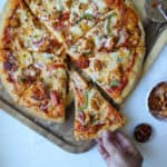 A hand reaches for a slice of Tandoori Turkey Pizza topped with cheese, green bell peppers, onions, and pieces of chicken on a wooden board. A pizza cutter and small bowl of toppings are nearby.