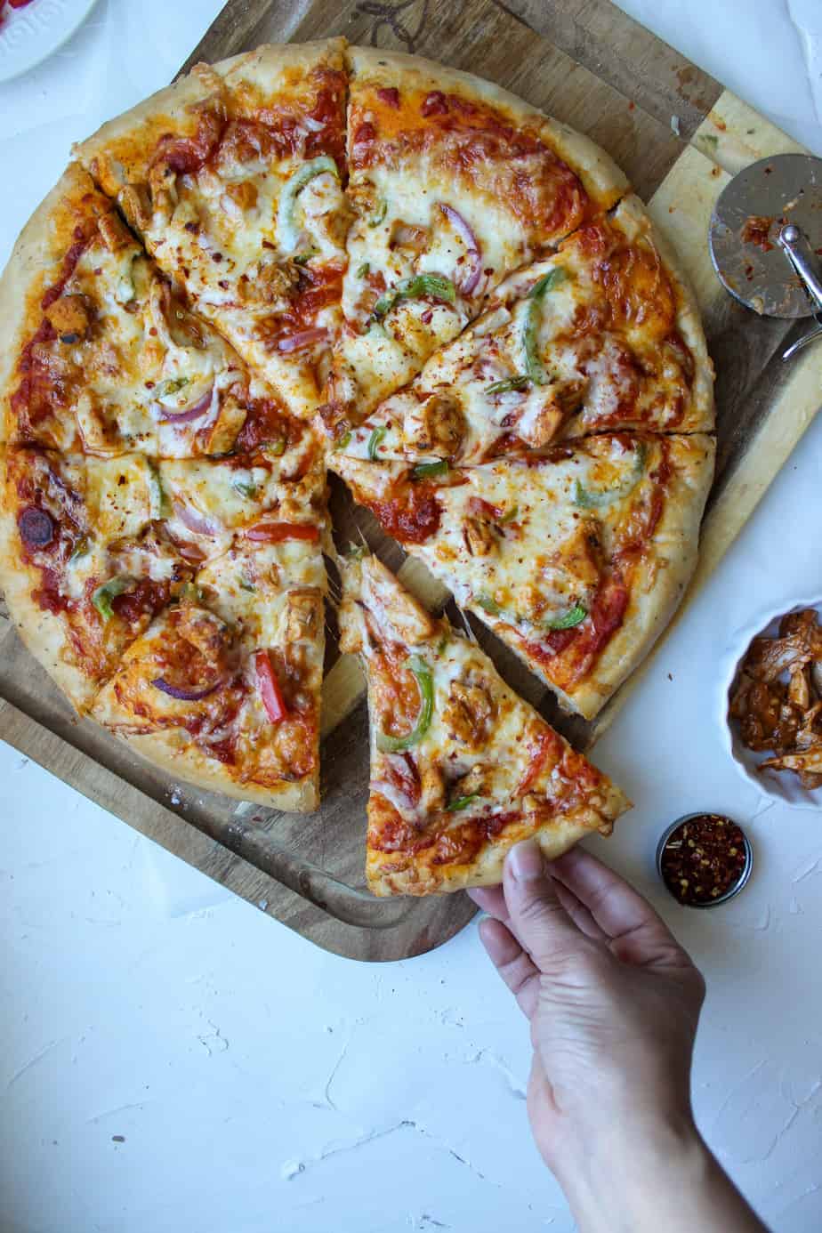 A hand reaches for a slice of Tandoori Turkey Pizza topped with cheese, green bell peppers, onions, and pieces of chicken on a wooden board. A pizza cutter and small bowl of toppings are nearby.
