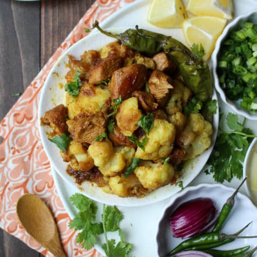 A bowl of Indian-style cauliflower and potato curry inspired by Pakistani cuisine, garnished with cilantro and surrounded by lemon wedges, green onions, chilies, red onion, and rice. A wooden spoon rests beside the bowl.
