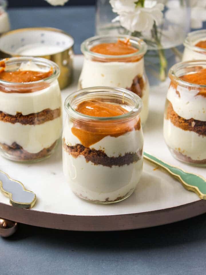 Small glass Cheesecake Jars filled with layers of cream, crumb, and caramel topping are arranged on a round marble board. A hand is about to scoop from one jar with a spoon. White flowers are visible in the background.
