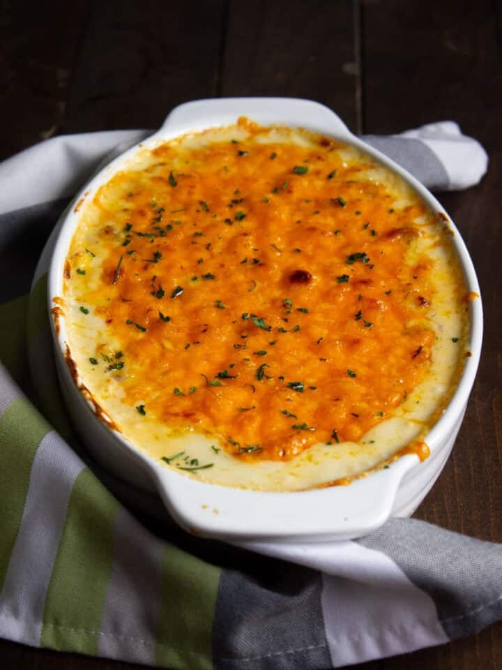 A white oval baking dish filled with baked chicken and cheese macaroni, topped with melted golden-brown cheese and sprinkled with herbs, sits on a striped green, white, and gray kitchen towel on a dark wooden surface.